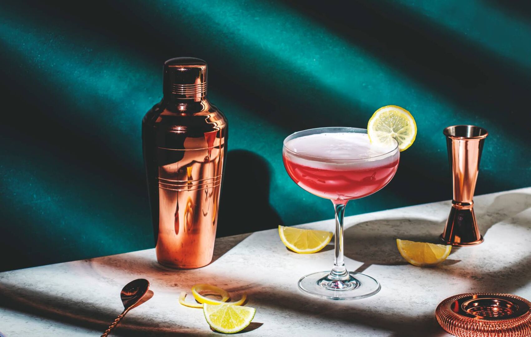 Ein elegantes Cocktailglas gefüllt mit Gin und einer garnierten Zitrone und einem Mixer aus Kupfer im Hintergrund auf einer weißen Tischplatte.