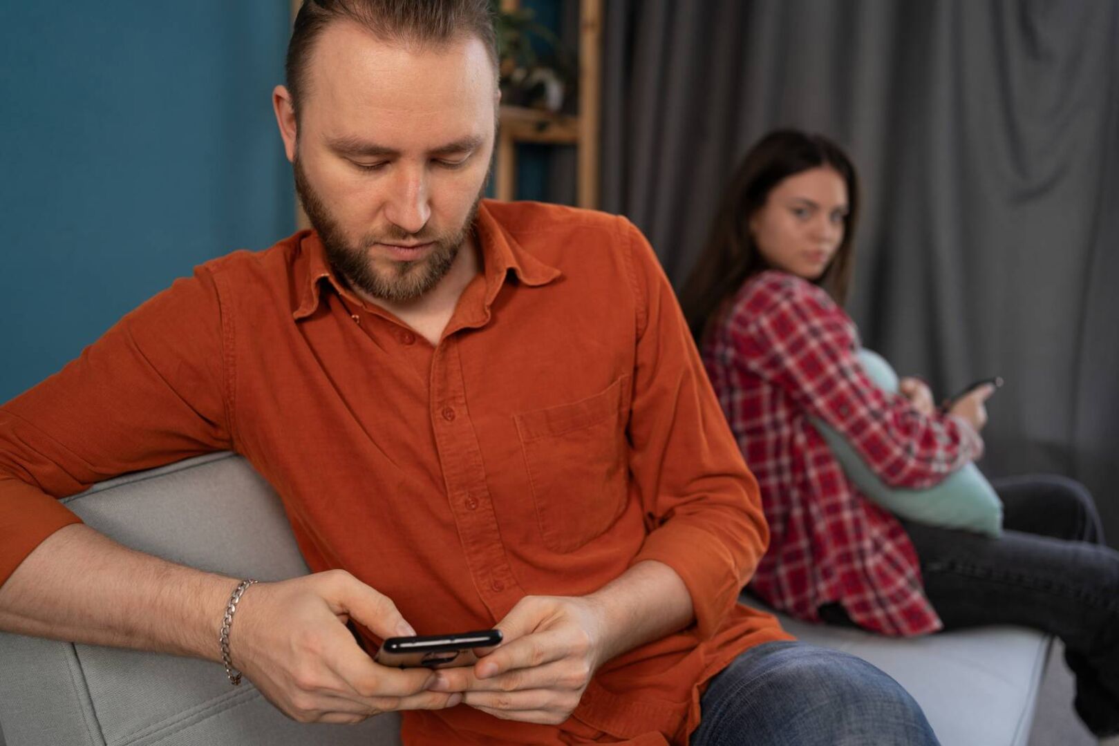 Ein Paar sitzt zusammen auf dem Sofa. Er hat sein Handy ausgepackt, während sie mit verschränkten Armen am anderen Ende des Sofas sitzt.