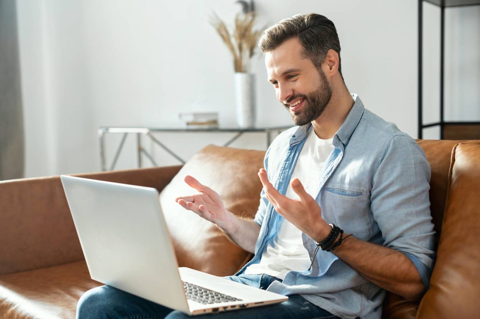 Junger Mann sitzt auf dem Sofa mit einem Laptop auf dem Schoß und führt ein Videotelefonat.