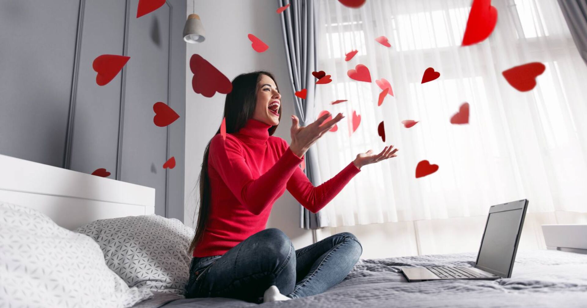 Eine junge Frau sitzt mit ihrem Laptop auf dem Bett. Um sie herum fliegen rote Papierherzen.