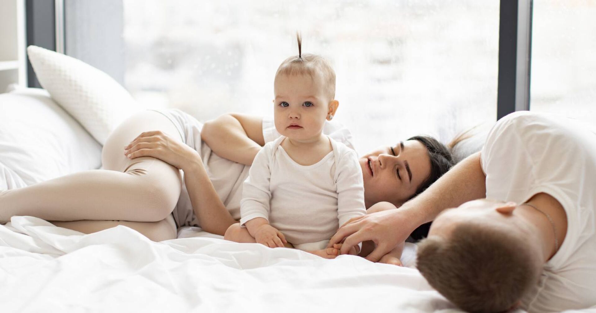 Paar mit Baby, das nicht schlafen will und kerzengerade auf dem Bett sitzt.