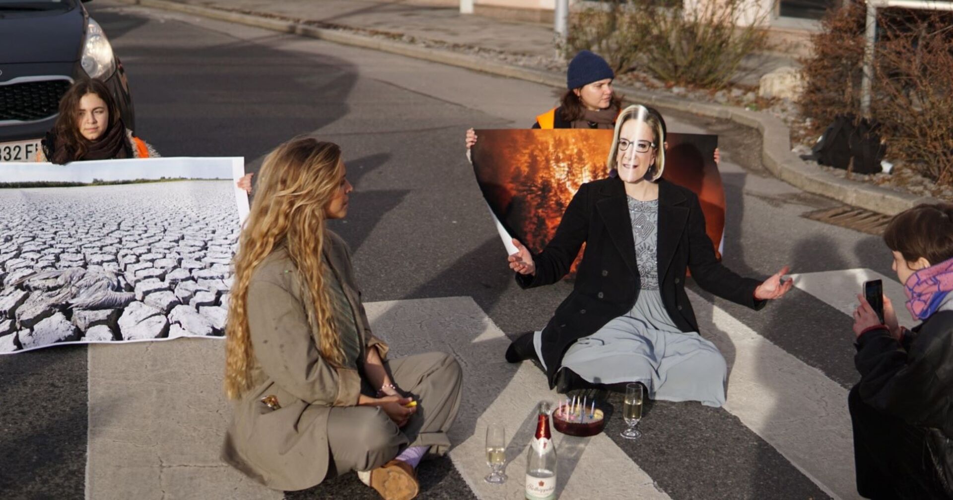 Aktivisten der letzten Generation sitzen auf einem Zebrastreifen. Sie halten Transparente, eine Person trägt eine Mikl-Leitner-Maske.