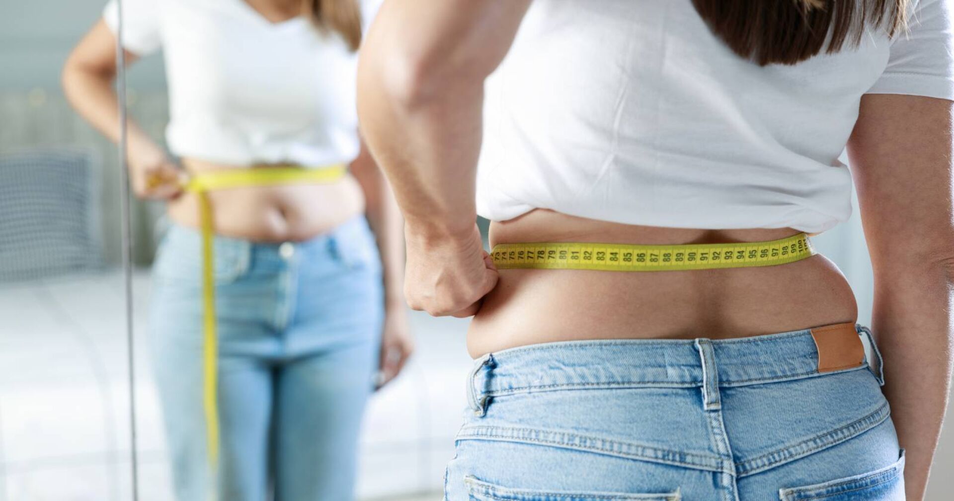 Frau vor einem Spiegel mit einem Maßband um die Taille zur Messung des Körperfettanteils