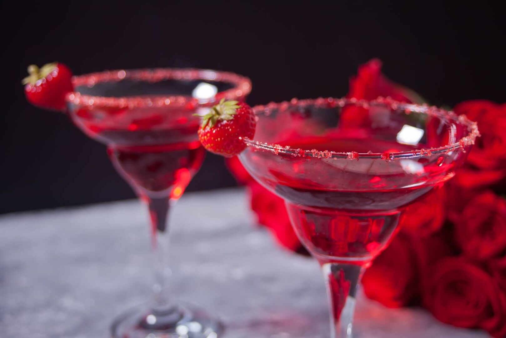 Zwei leuchtend rote, exotische alkoholische Getränke in transparenten Gläsern, begleitet von roten Rosen für ein romantisches Abendessen.