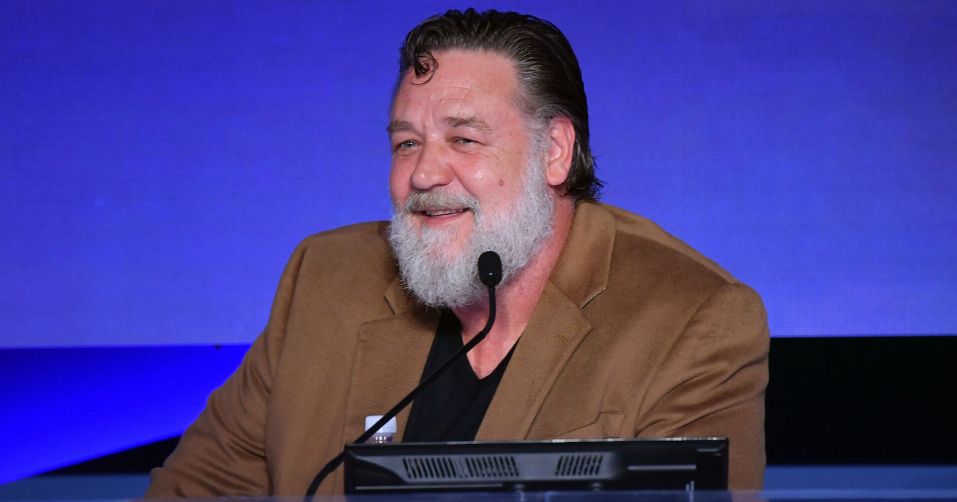 Russell Crowe in einem beigen Sakko bei einem Event. Er trägt einen Vollbart.