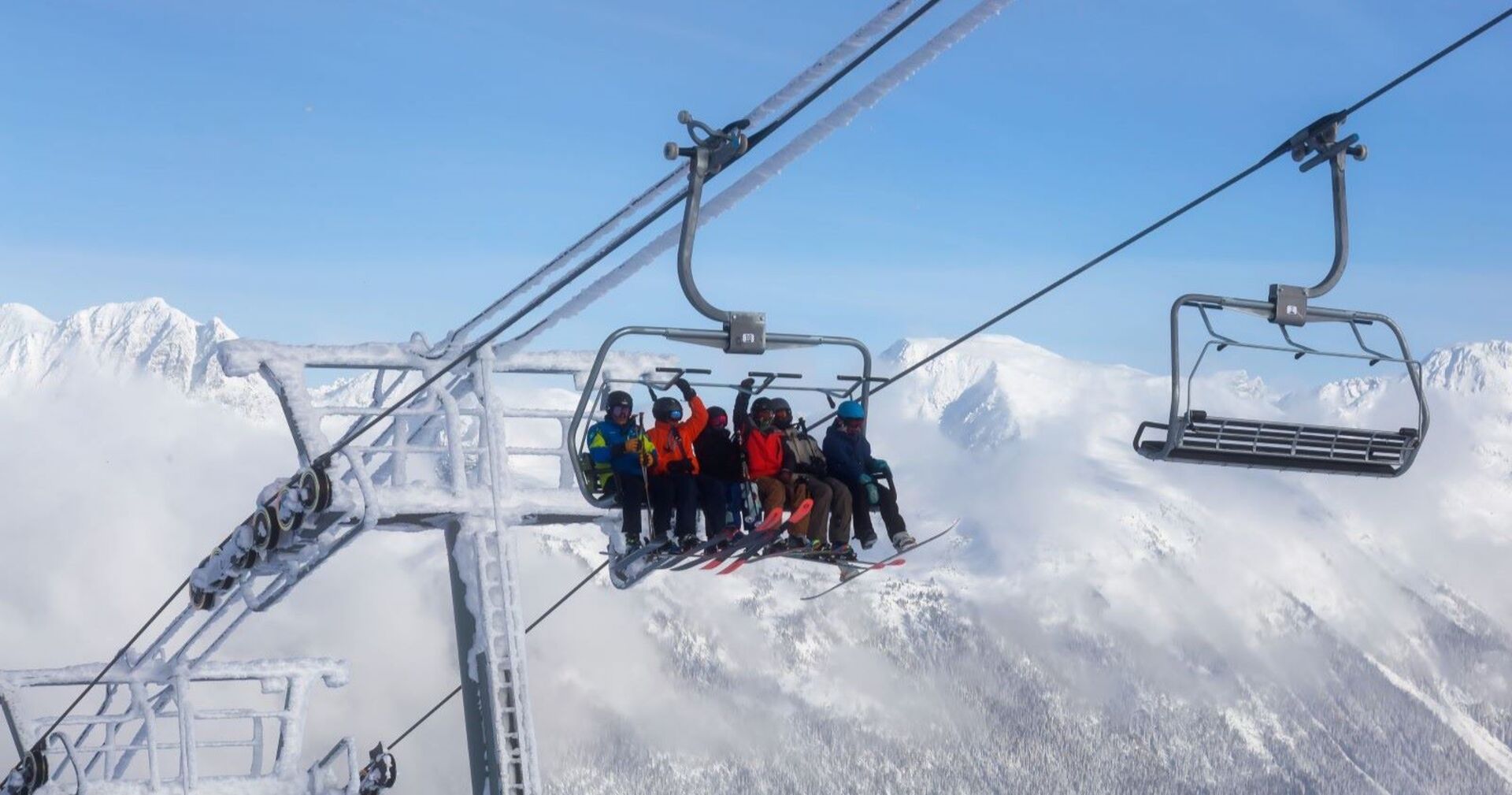 Ein Sessellift mit acht Skifahrern in der Luft, im Hintergrund eine schneebedeckte Gebirgskette