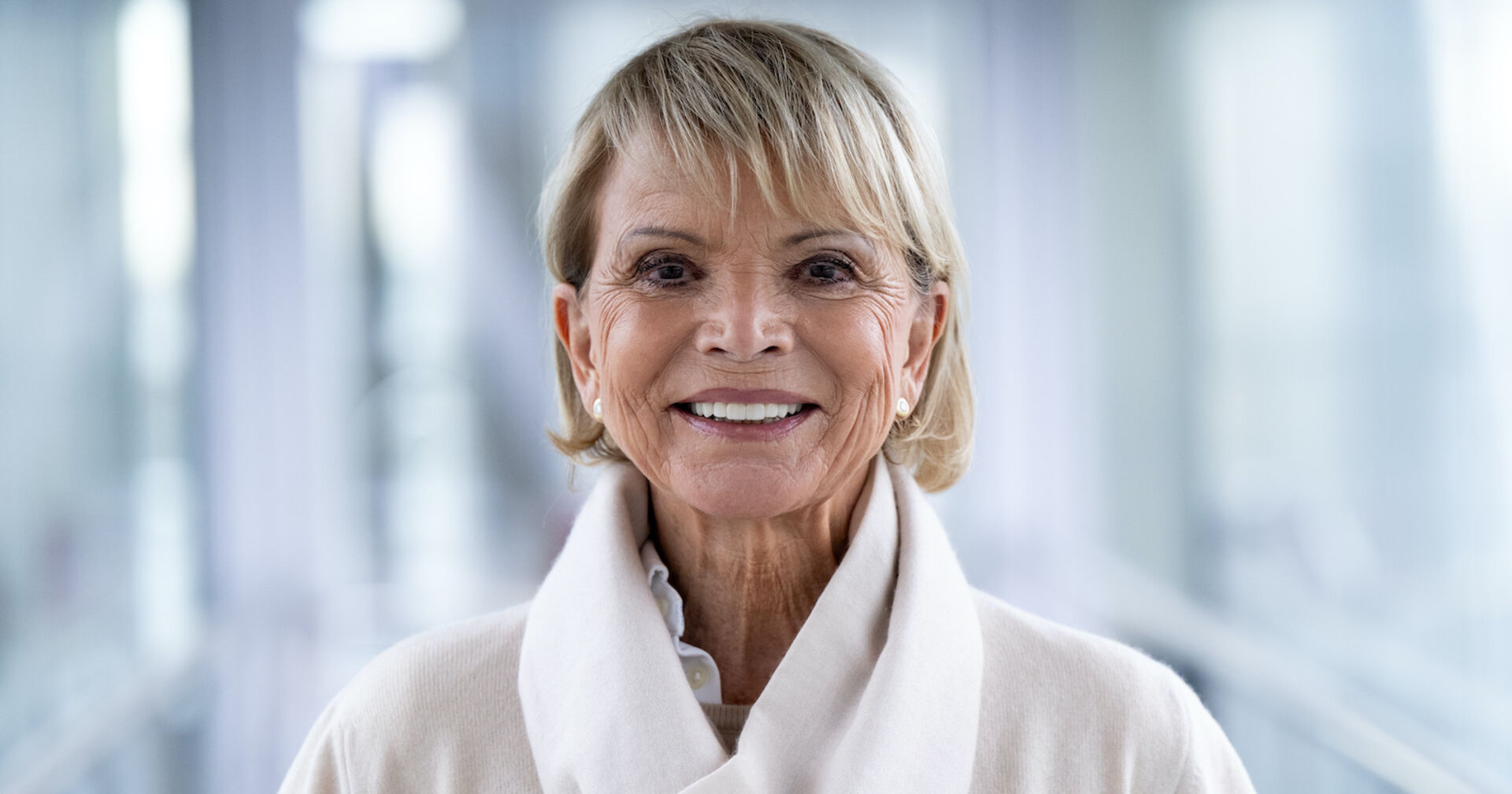 Uschi Glas mit einem weißen Schal und einem beigen Pullover. Sie lacht in die Kamera.