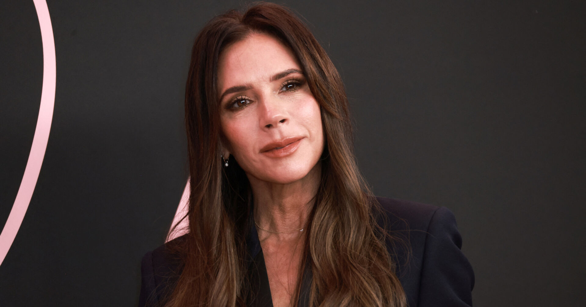 Victoria Beckham mit langen braunen Haaren und einem schwarzen Outfit vor einer schwarzen Fotowand.