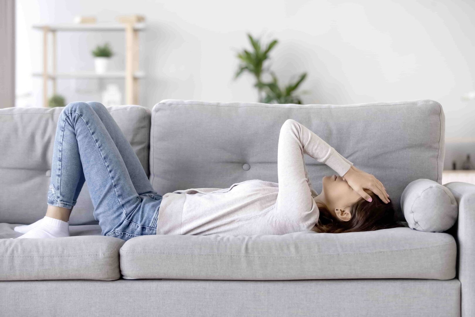 Eine Frau - mit weißen Pullover und einer blauen Jean bekleidet - liegt auf einer grauen Couch. Ihre Hände hat sich nachdenklich über ihr Gesicht gelegt.