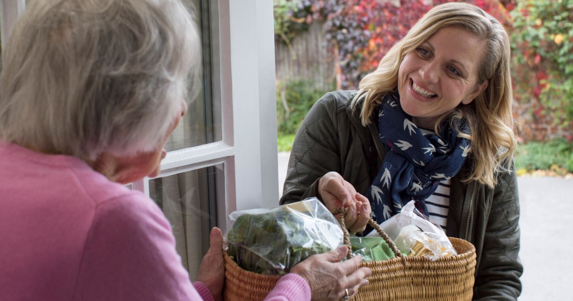 Frau überreicht älterer Dame einen Einkaufskorb an der Haustür