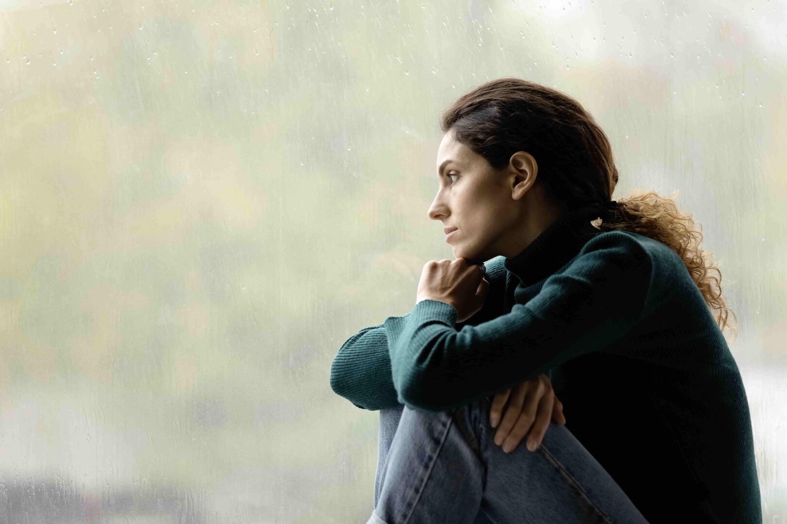 Eine Frau im mittleren Alter trägt einen grünen Pullover und sitzt am Fenster. Ihr Blick ist traurig und fragend.