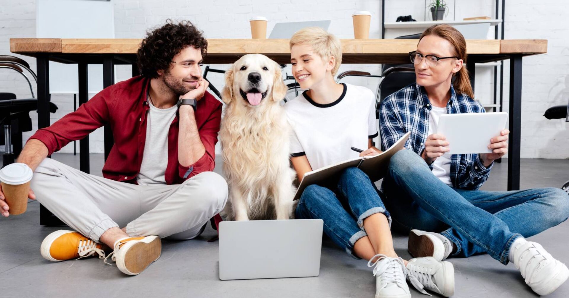 Drei Menschen sitzen zusammen mit einem Golden Retriever vor einem Laptop auf dem Büroboden und lachen zusammen.