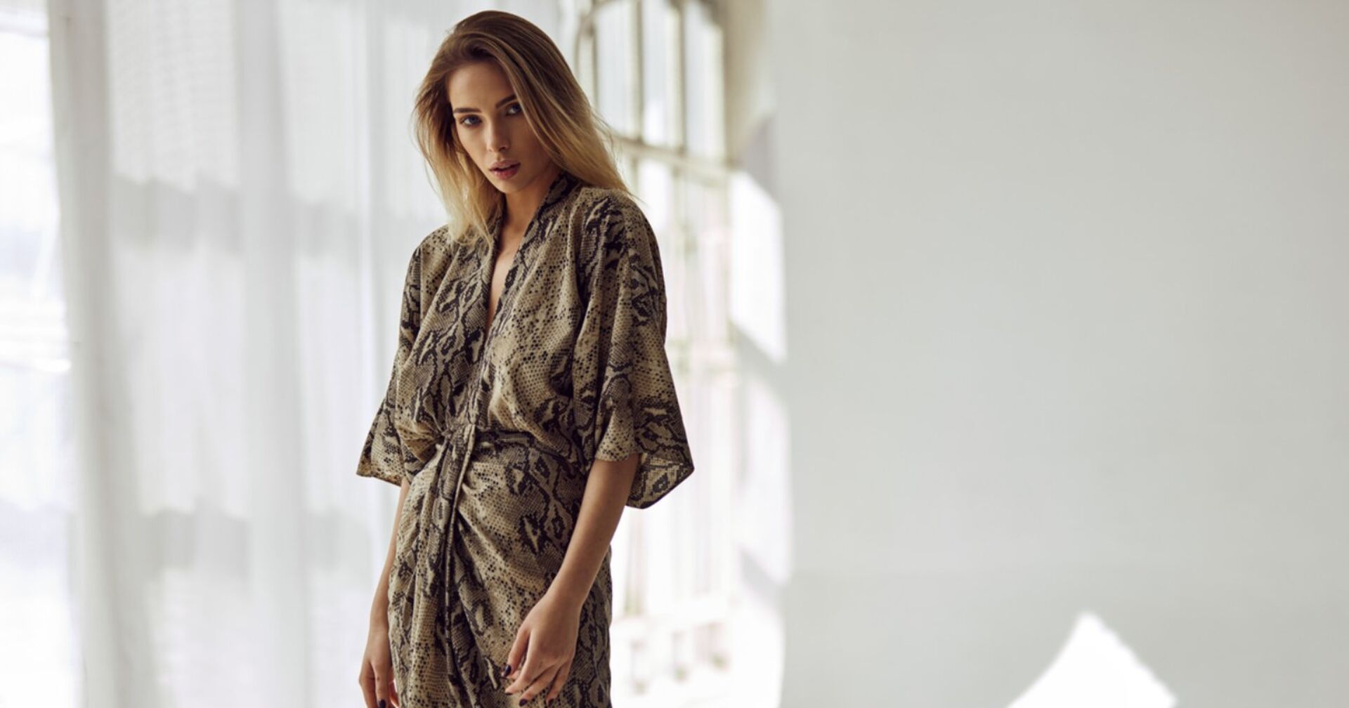 Eine Junge Frau mit braunen langen Haaren posiert in einem langen und luftigen beigen Kleid mit dunkelbraunem Muster.