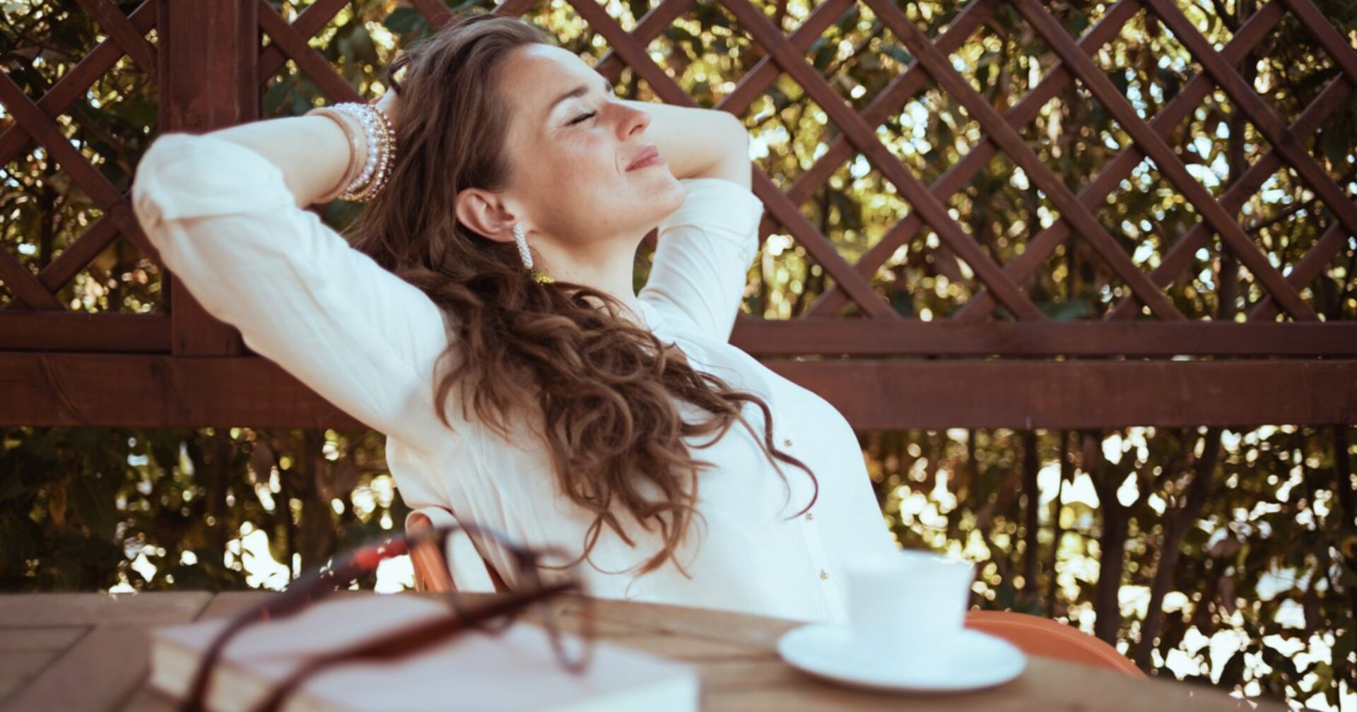 Ein junge Frau mit langen braunen Haaren, sitzt mit ihren Armen hinter dem Kopf verschränkt im Freien, hat ihre Augen zu und genießt die frische Luft. Vor ihr steht eine weiße Tasse auf dem Tisch sowie ein Buch und eine Brille.
