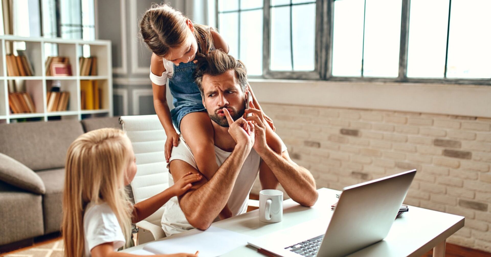 Ein junger Vater sitzt an seinem Schreibtisch vor einem Laptop, rechts neben ihm ein kleines blondes Mädchen, das mit Buntstiften malt und auf seinen Schultern sitzt ein kleines braunhaariges Mädchen | Credit: iStock.com/Valerii Apetroaiei