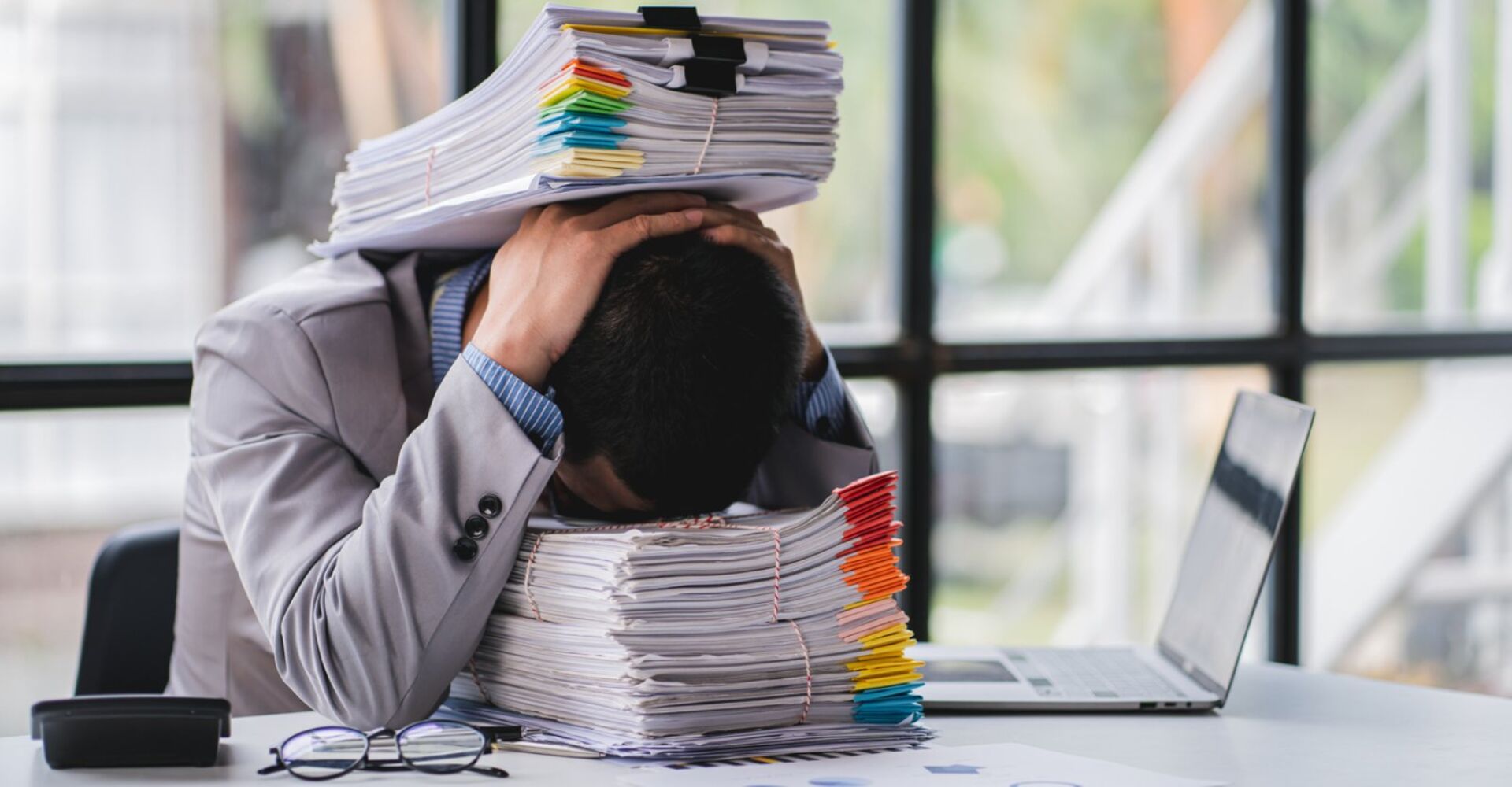 Ein Geschäftsmann sitzt an seinem Schreibtisch, sein Kopf auf einem Stapel Dokumente abgelegt und weitere Dokumente stapeln sich auf seinem Kopf.