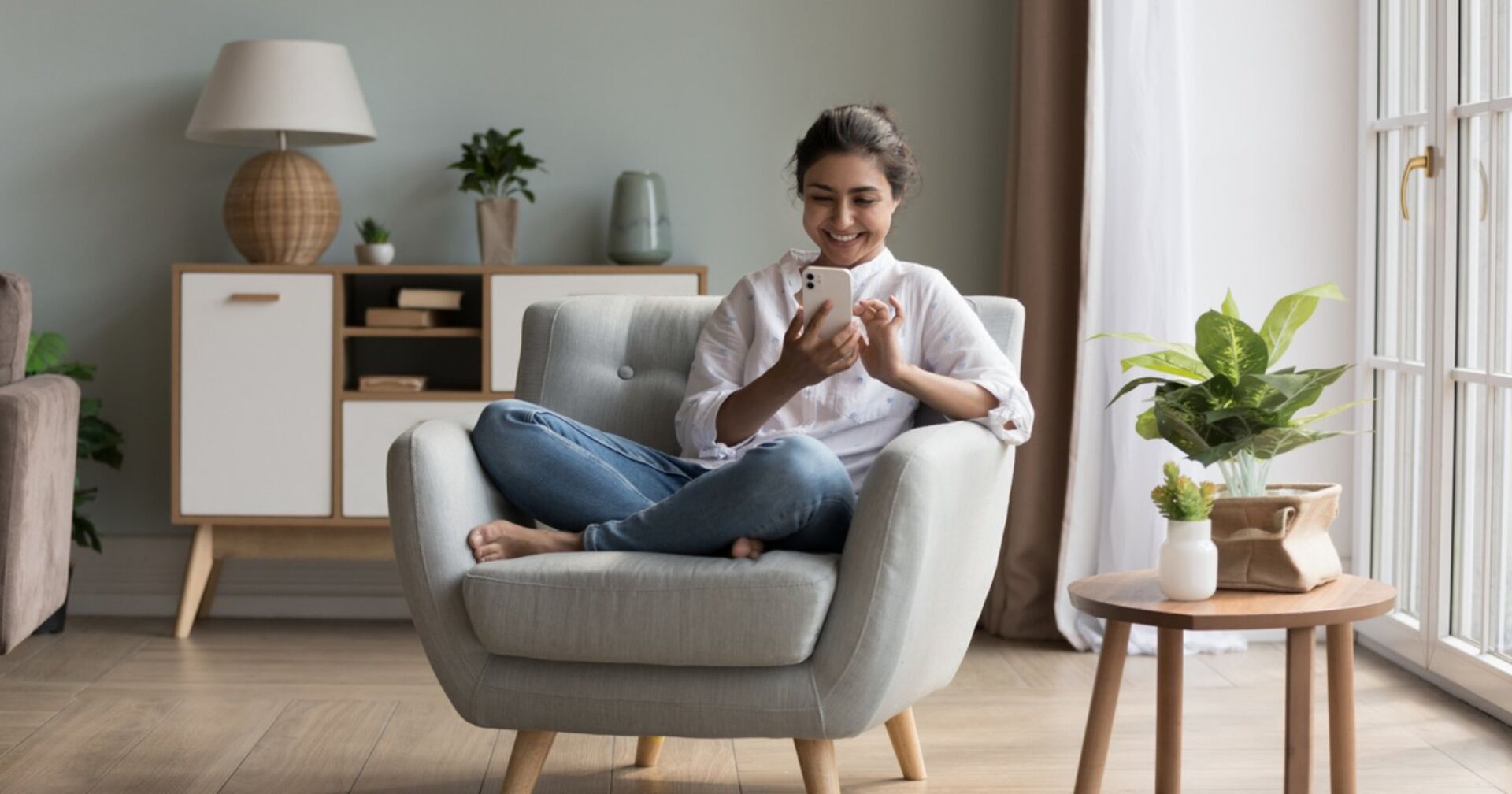 Eine Frau sitzt zuhause auf einem gemütlichen grauen Sessel und lacht in ihr Handy. Neben ihr befindet sich ein kleiner Holztisch mit einer Pflanze und hinter ihr ist eine Kommode zu sehen, auf der eine kleine Lampe sowie eine weitere Pflanze platziert sind.