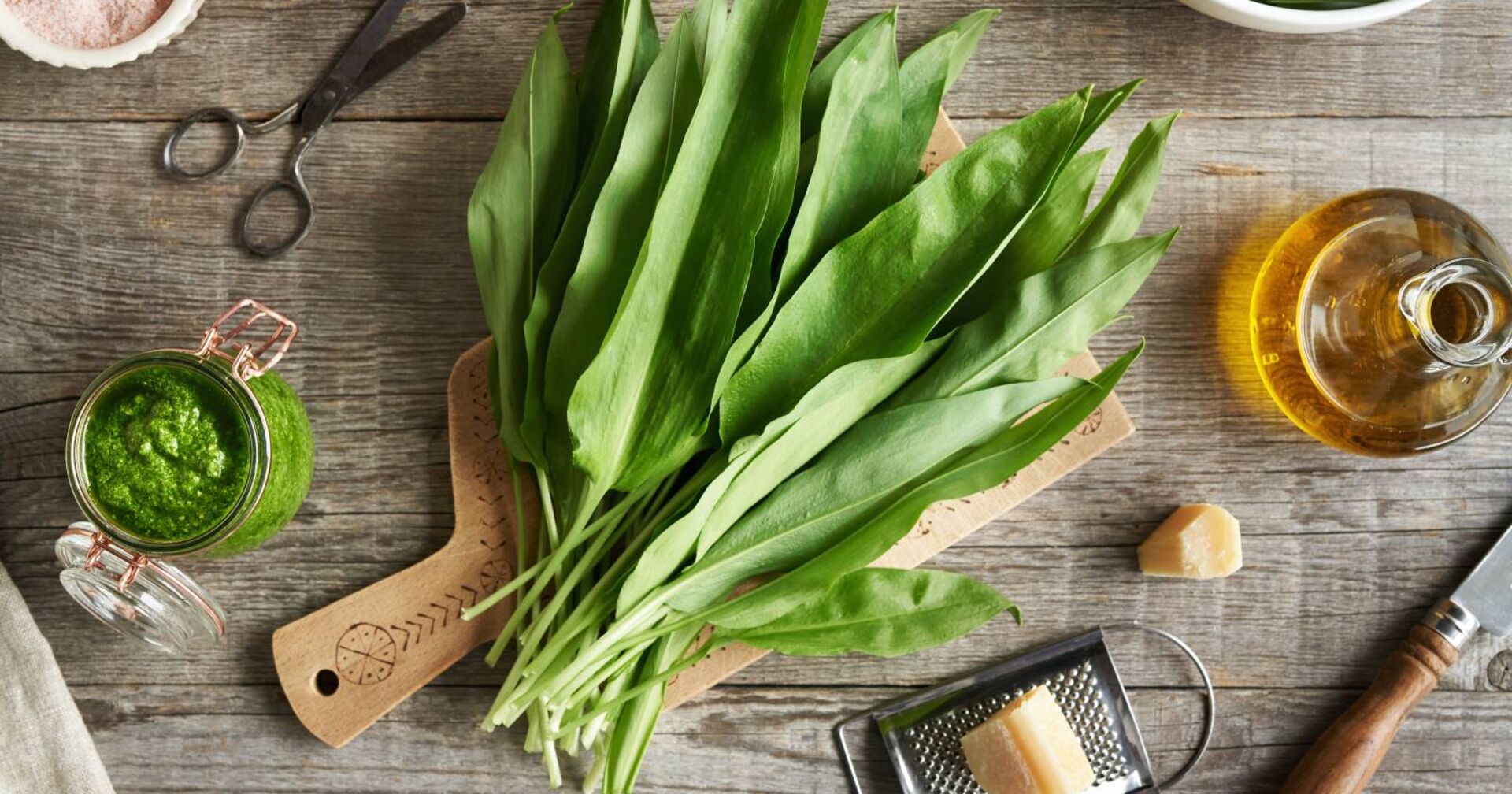 Ein Bund Bärlauchblätter auf einem Hackbrett umgeben von einer mit Öl befüllten Glasflasche sowie einer Käsereibe mit Parmesan