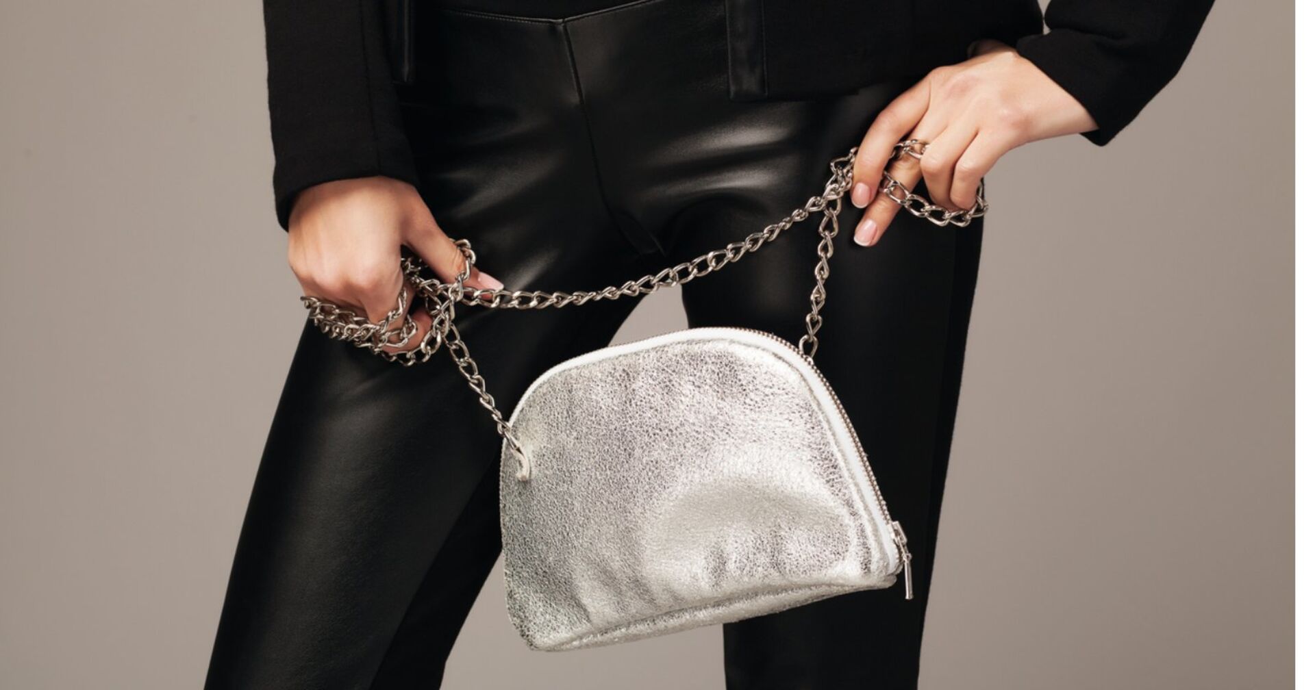 Frau hält silberne metallic Handtasche mit Riemen in Ketten-Optik in der Hand und trägt eine schwarze Lederhose sowie einen schwarzen Pullover.