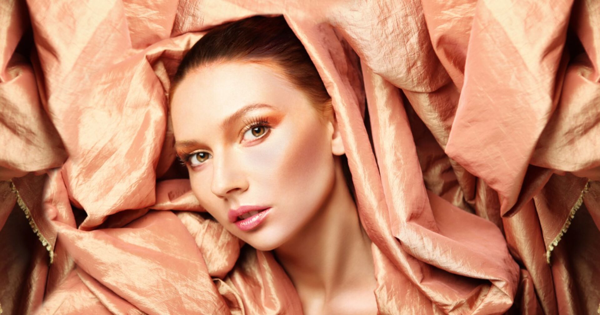 Frau mit peach-farbenem Makeup umgeben von metallischem Stoff in Pfirsichtönen