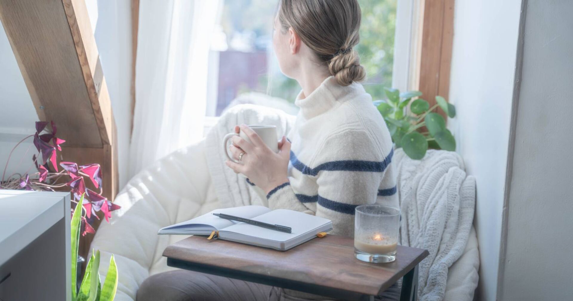 Junge Frau mit Blick aus dem Fenster und aufgeschlagenem Notizbuch auf dem Schreibtisch.