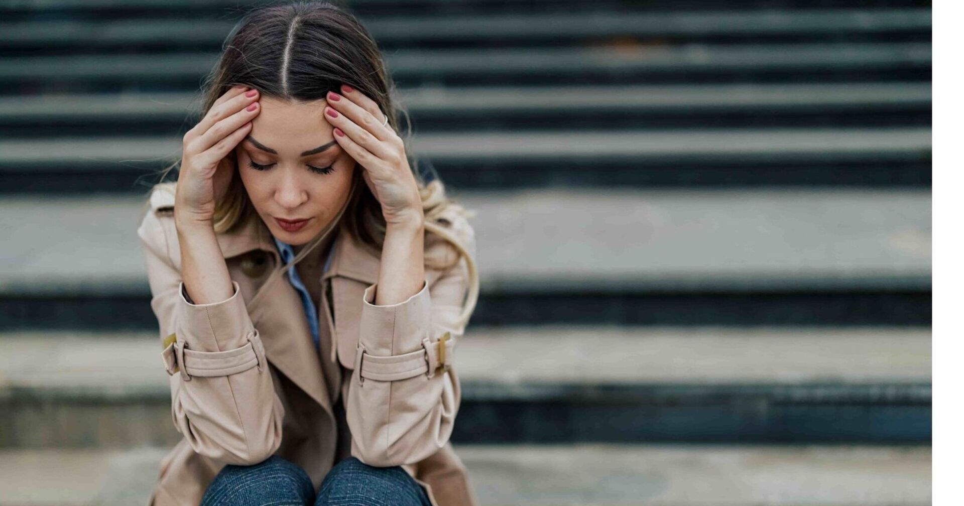 Eine Frau mit langen braunen Haaren sitzt Stufen. Ihre beiden Hände stützen ihren Kopf, ihr Gesicht ist traurig und nachdenklich. Ihre Augen sind geschlossen.