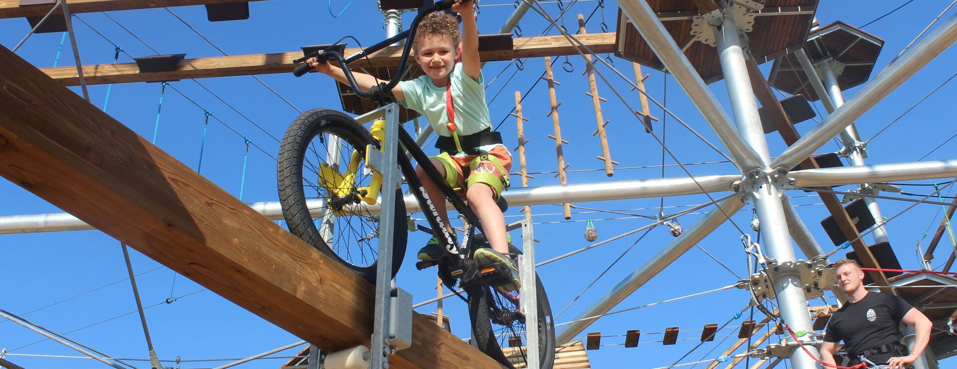 Ein junge balanciert mit einem Mountainbike auf einem Holzbalken. Er trägt ein grünes T-Shirt und eine kurze, gelbe Hose. Er ist mit Seilen und einem Gurt gesichert. Im Hintergrund sieht man den Klettergarten des Naturparks Ikuna mit Brücken, Seilen und anderen Hindernissen.
