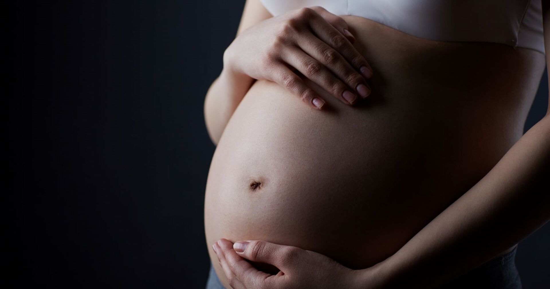 Eine Frau hält zärtlich ihren schwangeren Bauch mit beiden Händen, die Grundstimmung ist düster.