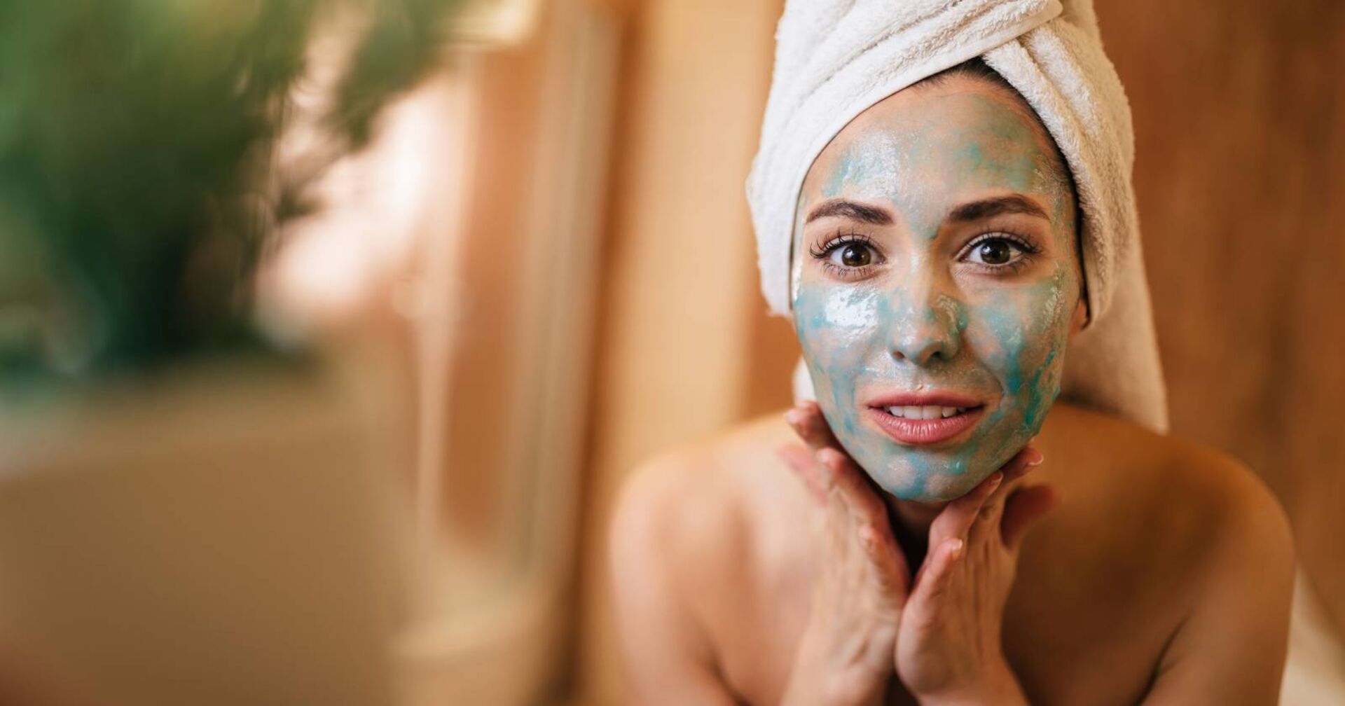 Junge Frau mit grüner Gesichtsmaske, die sich im Spiegel betrachtet.