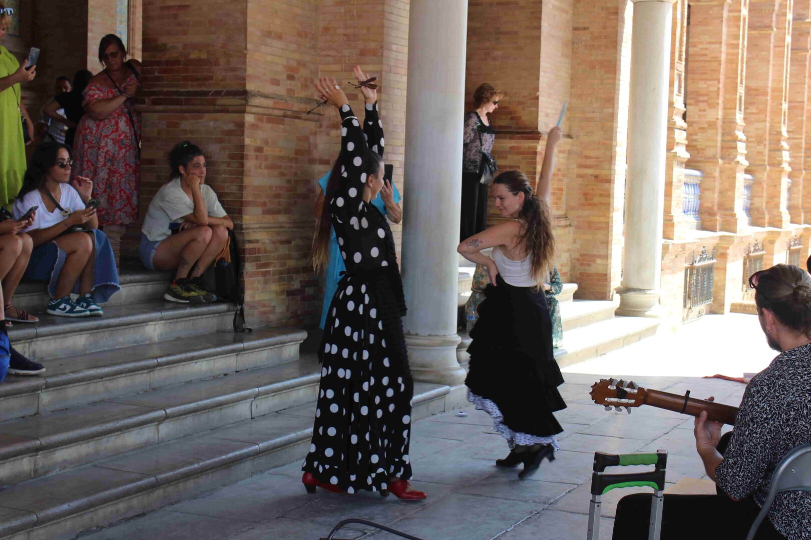 Im Schatten des Plaza De España tanzen zwei Spanierinnen Flamenco. Eine der Damen trägt ein langes schwarzes Kleid mit weißen Punkten. Die andere einen schwarzen Rock und ein weißen, kurzärmeliges Hemd. Begleitet werden sie dabei von einen Gitarrenspieler (links im Bild). Rechts davon sitzen Touristen, die den Klängen lauschen.