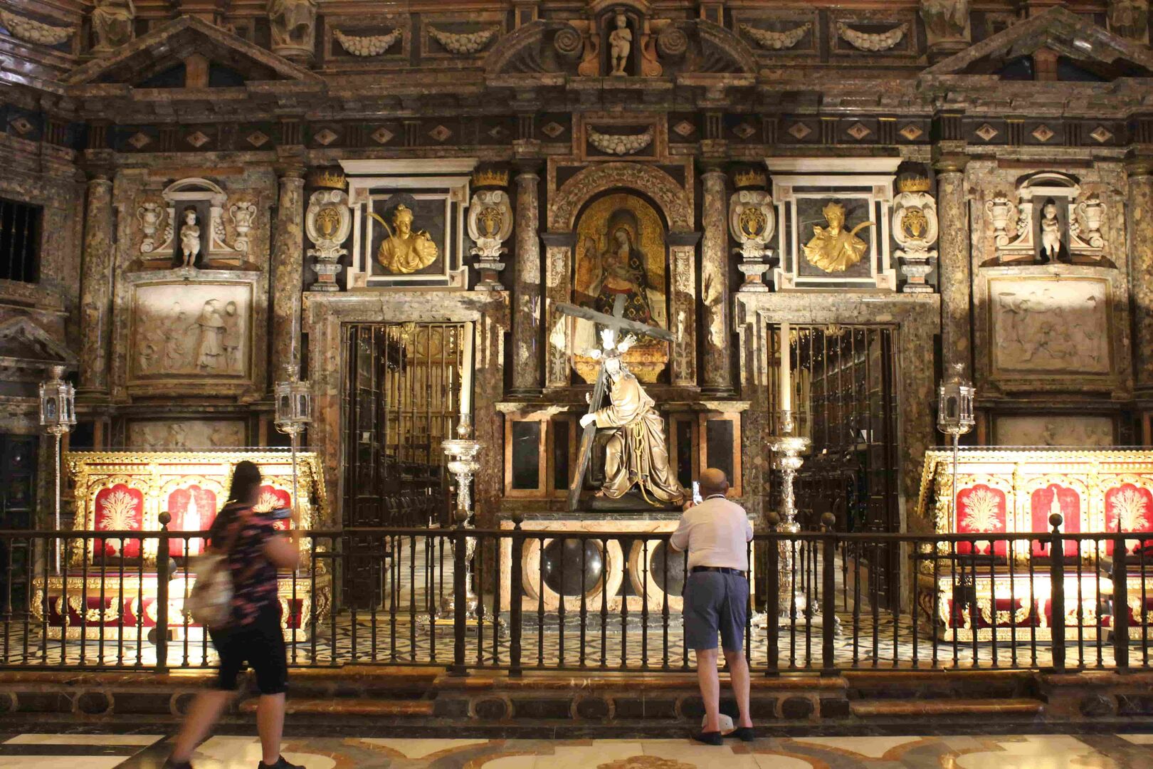 in Mann mit Glatze und kurzen, blauen Hausen steht vor einem Seit und kurzer Hose in der Kathedrale von Sevilla. Zu sehen ist Jesus, der sein Kreuz trägt und ein mit Gold verzierter Altarraum.