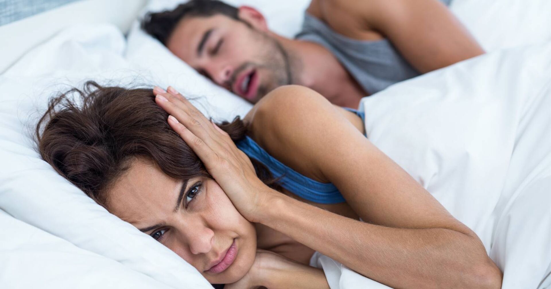 Junge Frau, die wach neben ihrem Partner im Bett liegt und sich die Ohren zuhält