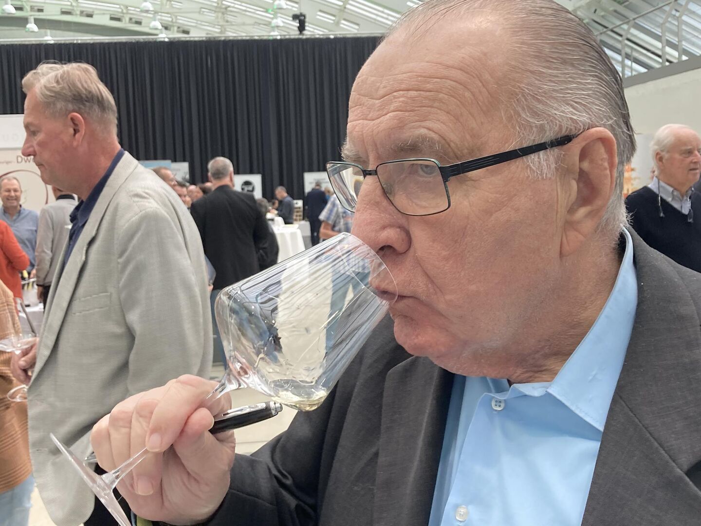 Ein Mann mit Brille und Halbglatze  - mit blauem Hemd und Sakko bekleidet - verkostet einen Weißwein. In der Hand hält er einen Bleistift und ein Glas mit Weißwein. Im Hintergrund sieht man andere Weinfreunde kosten und diskutieren.