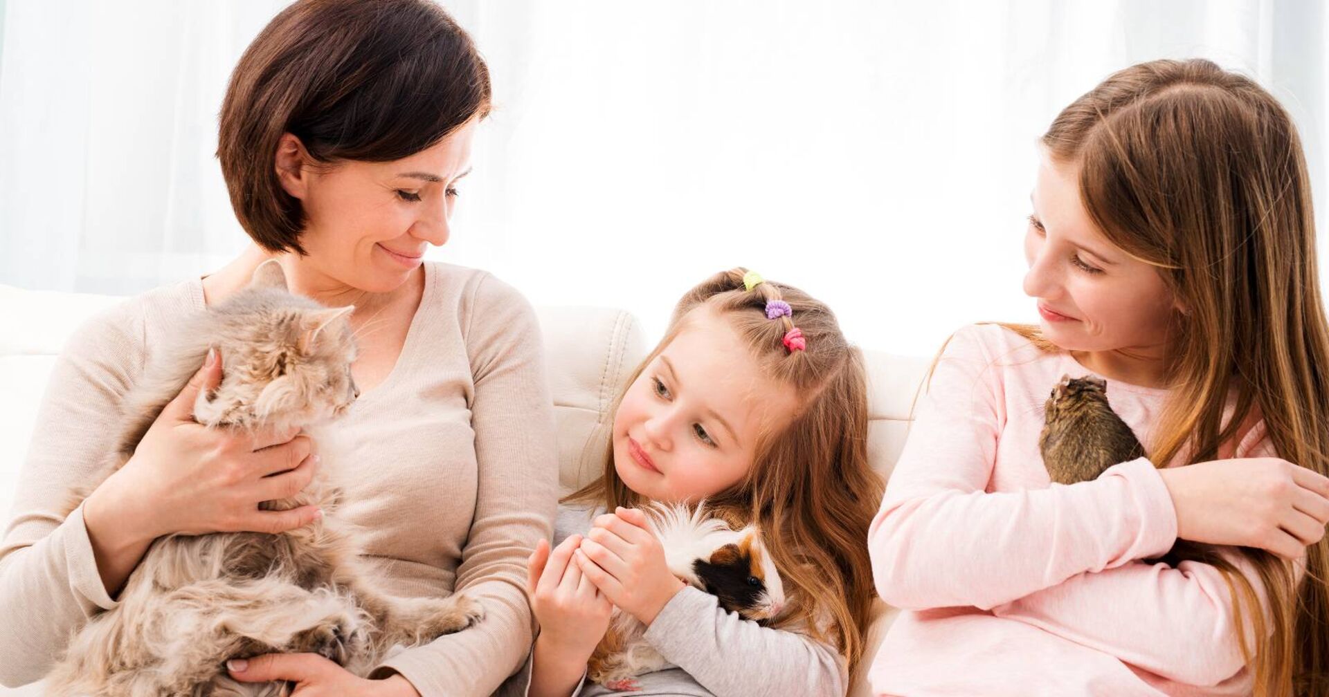 Eine Mutter sitzt mit ihren beiden Töchtern auf dem Sofa. Jede hält ein anderes Haustier auf dem Arm.
