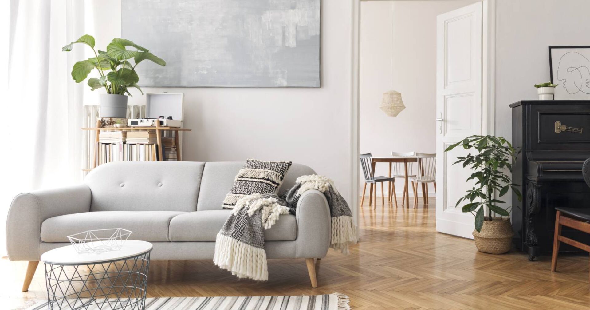 Wohnzimmer mit hellgrauer Couch, einem dunklen Klavier und viel Tageslicht, dass durch die Fenster des Zimmers hineinkommt.