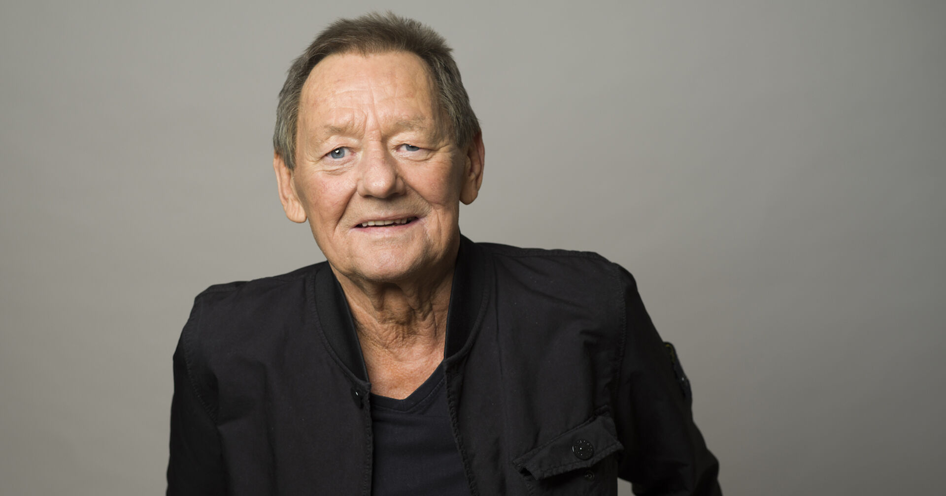 Wolfgang Ambros mit einem schwarzen Outfit vor einer grauen Fotowand.