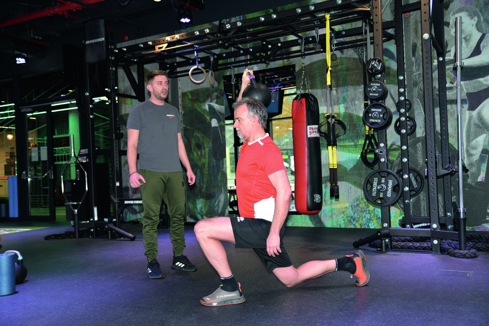 Mann hebt eine Kettle-Ball über den Kopf und ein Trainer beobachtet ihn