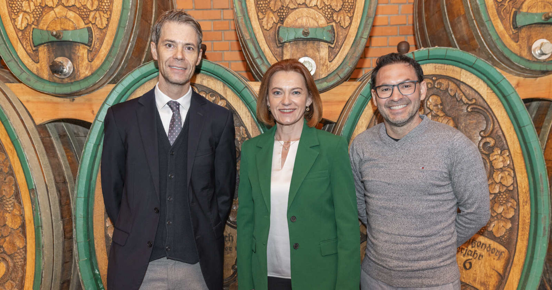 Zwei Männer und eine Frau vor braunen Weinfässern