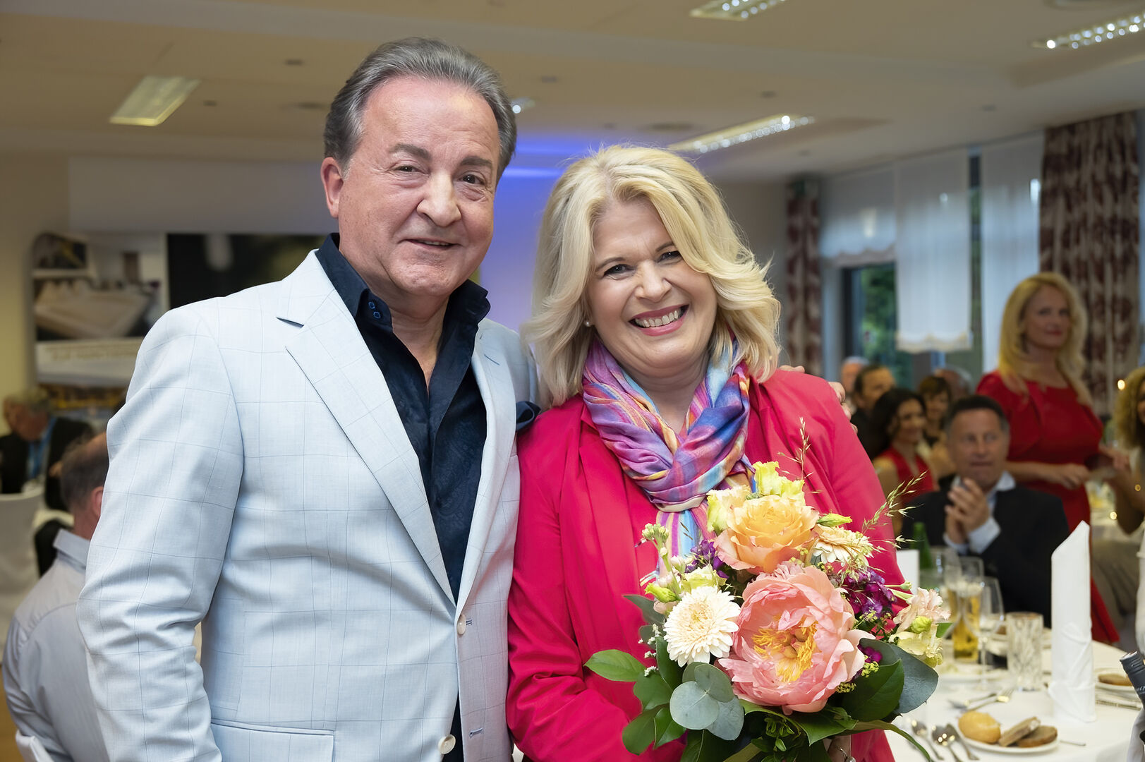 Ein Mann in blauem Sakko steht neben einer Frau, die Blumen in der Hand hält