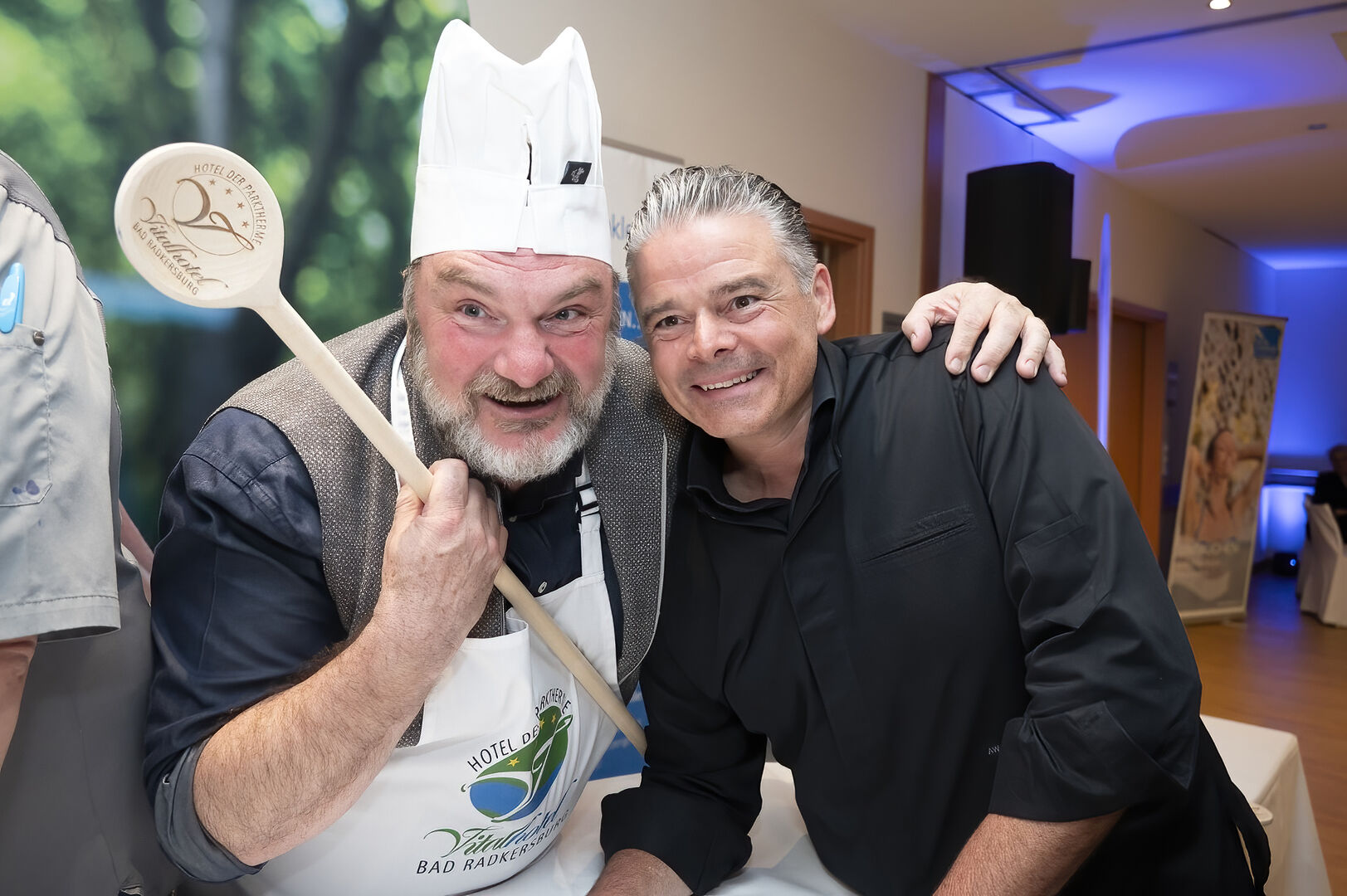 Ein Mann legt seinen Arm um einen anderen und hält in der anderen Hand einen Kochlöffel