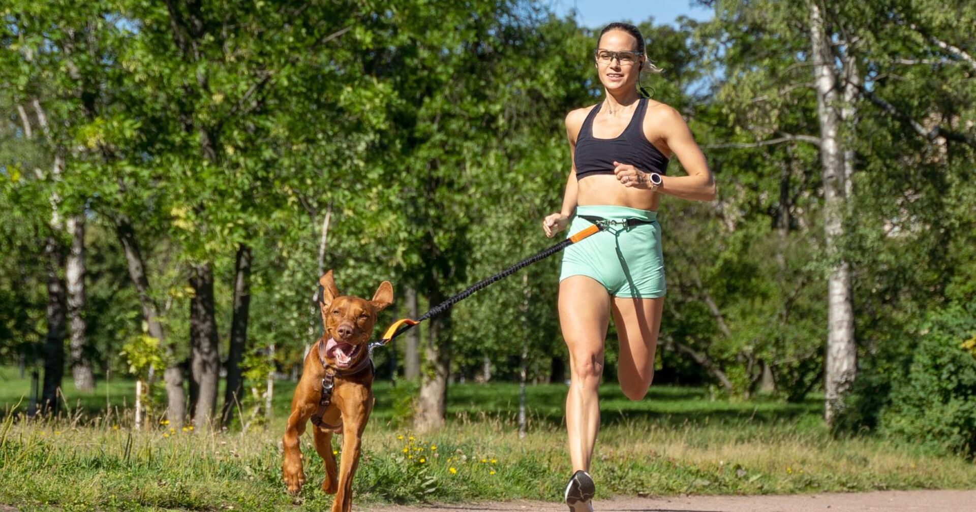 Junge Frau mit schwarzem Top, türkisfarbenen Shorts und Hund mit Canicross-Geschirr um die Hüfte