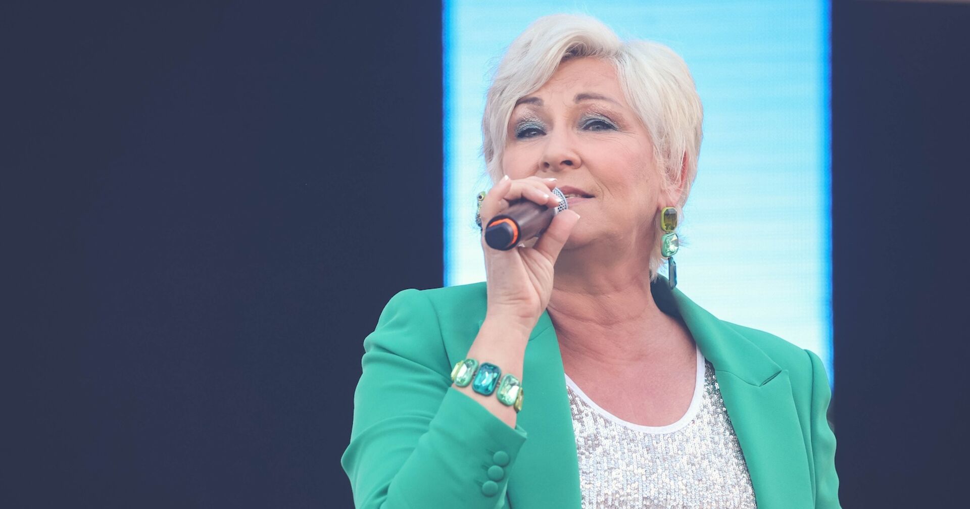 Claudia Jung mit einem türkisen Blazer, einem Mikrofon und kurzen blonden Haaren auf der Bühne.