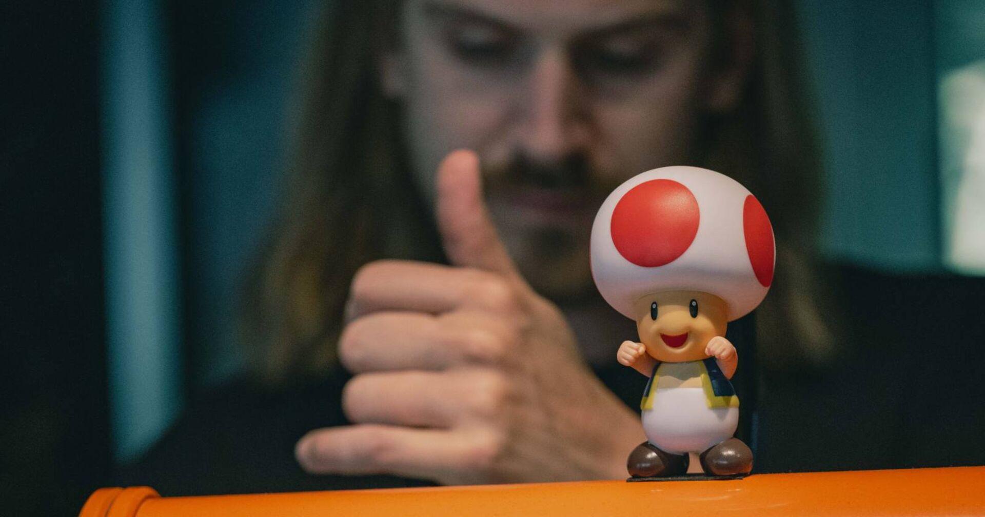 Das Bild zeigt einen Mann im Hintergrund, der anscheinend mit seinem Daumen nach oben zeigt, wahrscheinlich als Zeichen von Zustimmung oder Begeisterung. Im Vordergrund des Bildes steht eine Figur, die einem sehr bekannten Charakter aus einem populären Videospielfranchise.