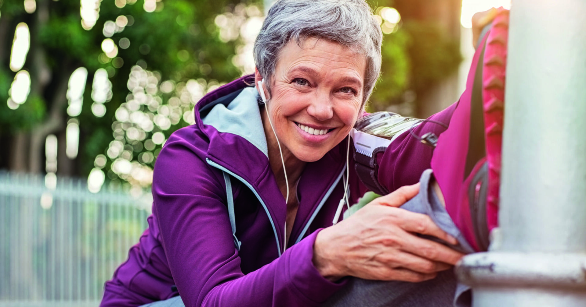 Eine ältere Frau - mit kurzen, grauen Haaren - streckt ihren Fuß an eine Säule. Sie trägt einen lila Trainingsanzug und dazu passende Turnschuhe.