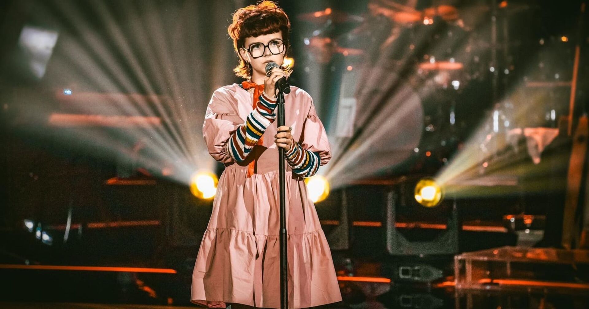 Victoria Knogler auf der steht auf der Bühne von &quot;The voice kids.&quot; Die Sängerin trägt ein rosa Kleid und einen Pullover mit bunten Querstreifen. Ihre braunen Haare sind kurz geschnitten, ihre Augen sieht man durch eine viereckige Brille. In der Hand hält sie ein Mikrofon.