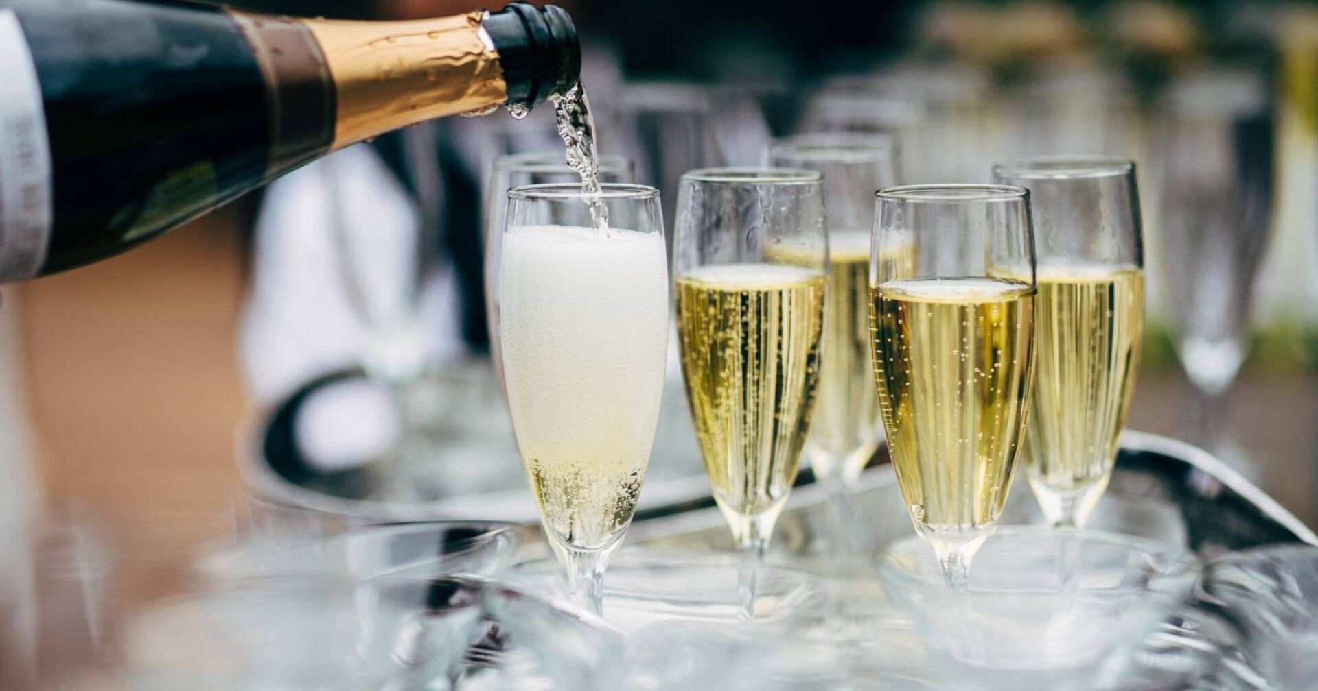 Mehrere Proseccogläser auf einem Tablett, im Vordergrund ein Glas, das gerade mit Prosecco gefüllt wird.