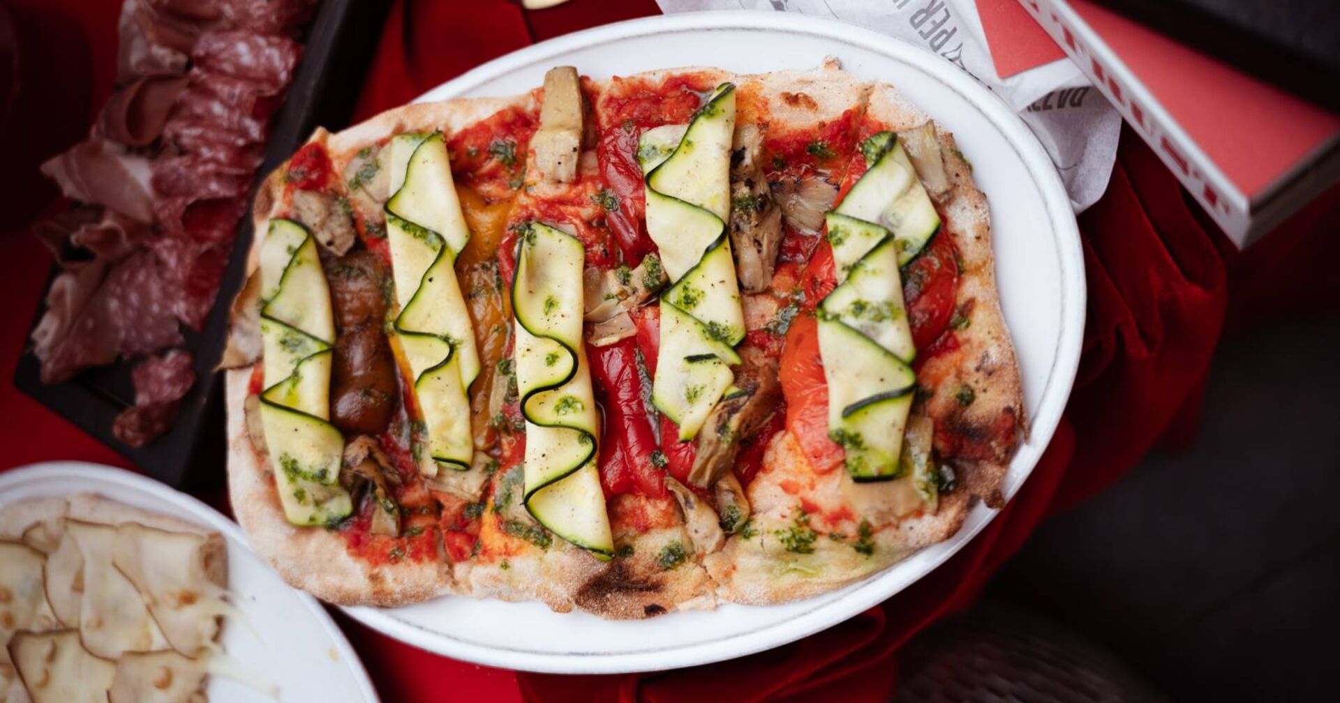 Zucchini und gegrillter Paprika auf einer römischen Pinsa. Im Hintergrund ist auf Tellern italienische Antipasti zu sehen.