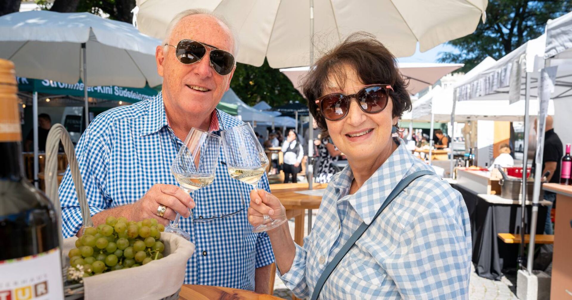 Zwei Menschen, die sich mit einem Glas Weißwein zuprosten und dabei lächelnd direkt in die Kamera schauen.