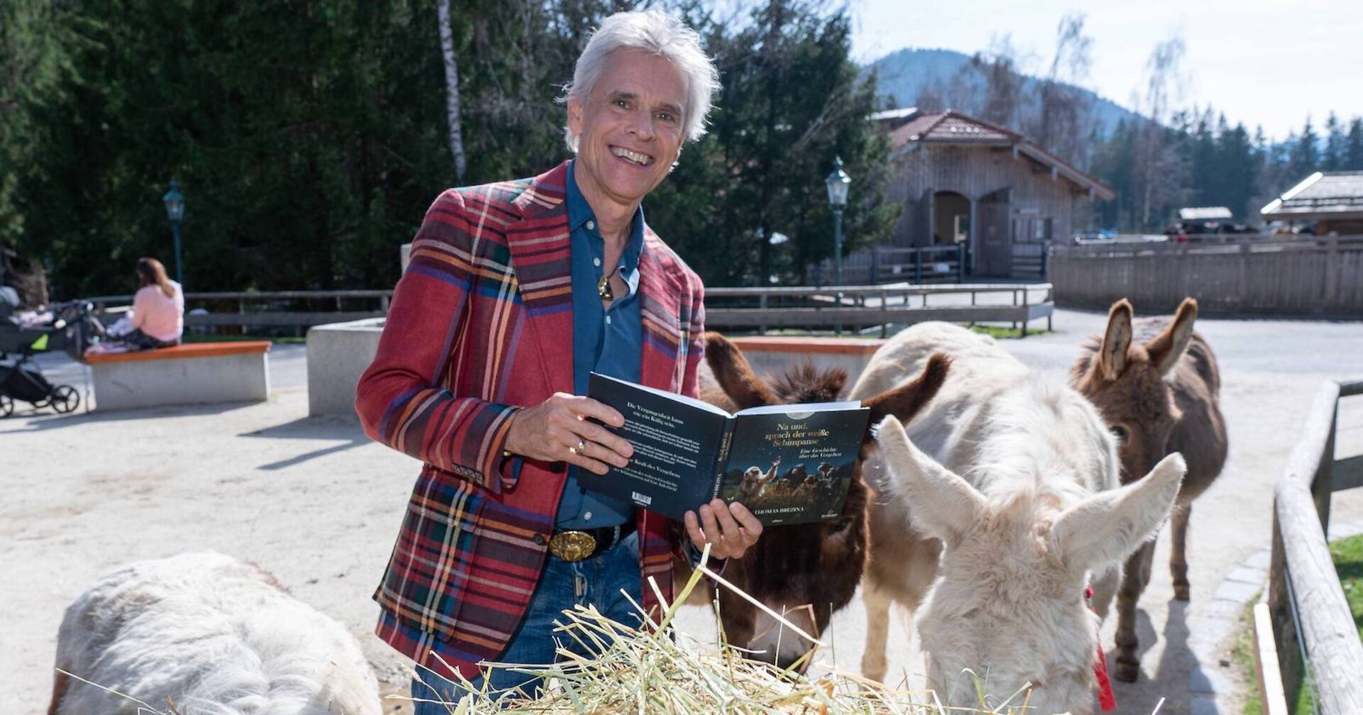 Thomas Brezina mit einem Buch, einem karierten Sakko und umgeben von Eseln.