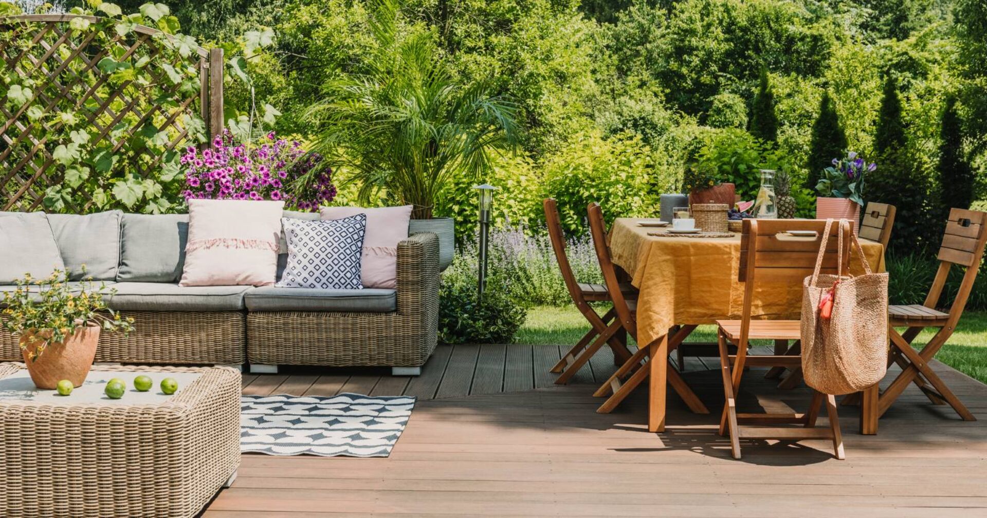 Blick in einen Garten mit Holztisch und Sesseln. Im Hintergrund ein Rattansofa.